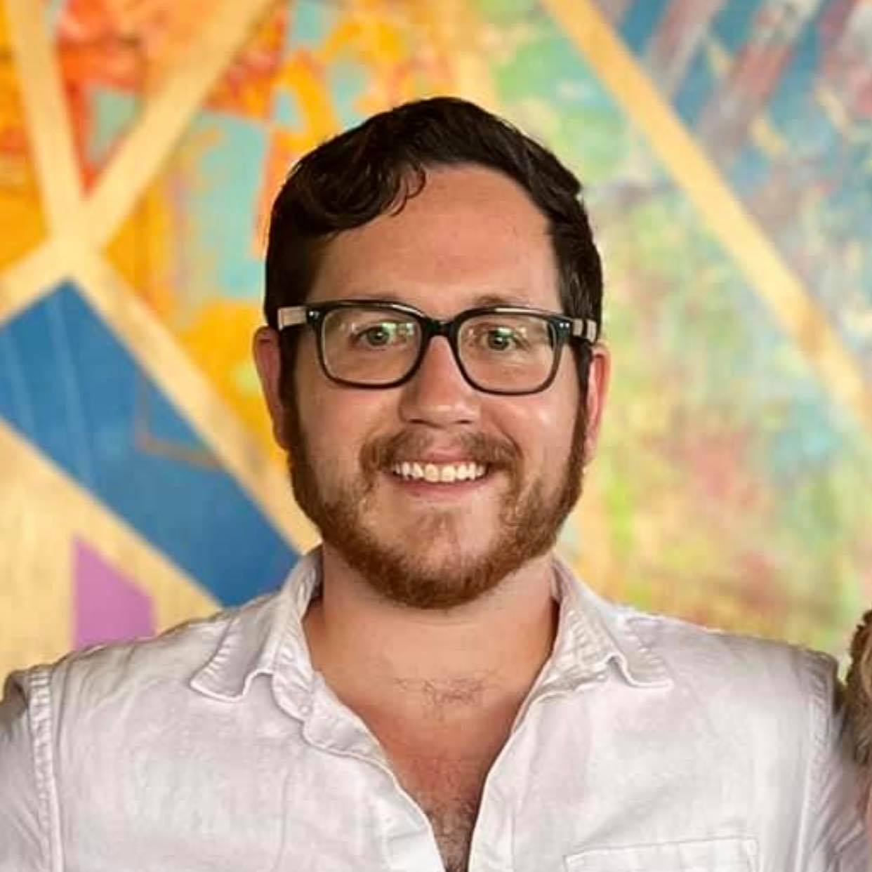 Isaac wears a white button up and black glasses smiling in front of a colorful background