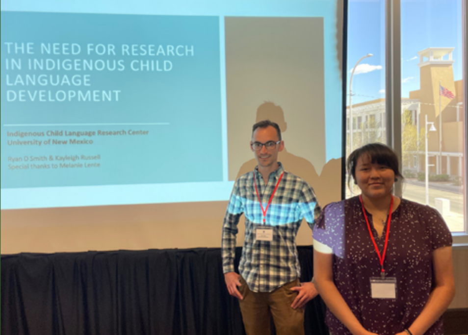 young man and young woman standing in front of a presentation slide which reads 'the need for research in Indigenous child language development'